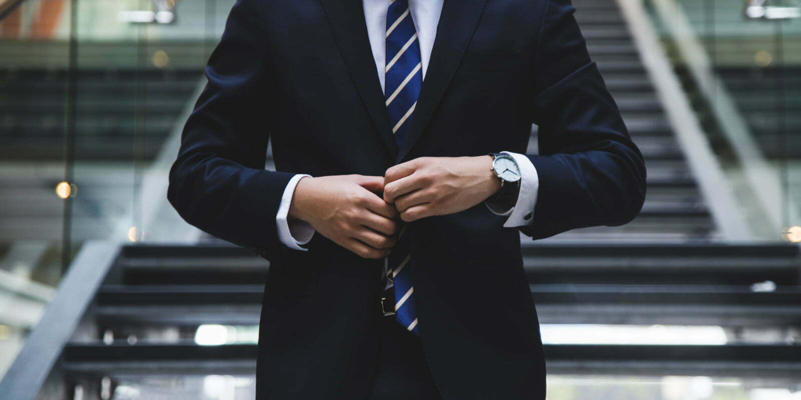 A close-up of a person standing at the bottom of a set of stairs fastening a dark blue single breasted suit.